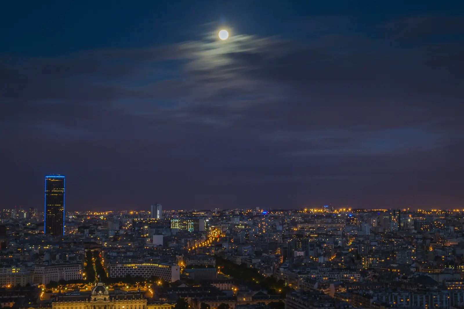 A moonlit city roofline viewed under a darkening evening sky.